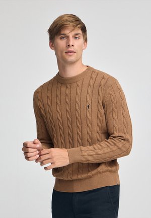 Bruine kabelgebreide trui met een ronde hals en geribde zoom. Bevat een subtiele logo-detail op de borst. Gedragen met een donkere broek.