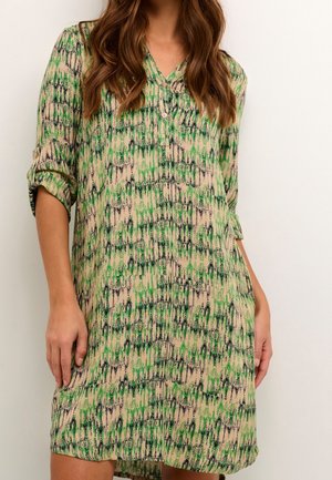 Femme portant une robe beige mi-longue avec un motif abstrait vert et noir et des manches retroussées, debout devant un fond uni.