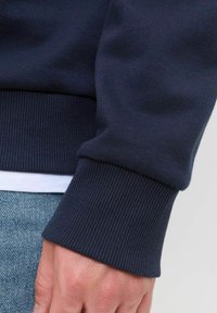 Marineblaue Sweatshirt mit gerippten Bündchen und strukturiertem Finish, aus weichem Stoff gefertigt, neben einem weißen Hemdsaum und Denim gezeigt.