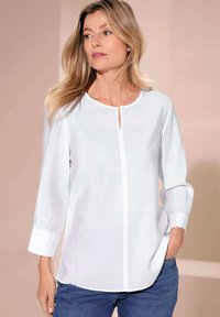 Blusa bianca con maniche 3/4, scollatura rotonda, dettaglio con bottoni a chiave e bordo a contrasto. Tessuto morbido e leggero, abbinato a jeans blu.