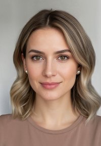 Donna con capelli biondi ondulati alla lunghezza delle spalle, che indossa una camicia marrone chiaro. Ha un trucco naturale e orecchini di perle bianche, su uno sfondo neutro.