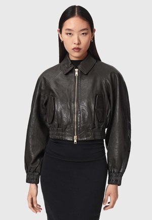 Jonge vrouw die een zwarte cropped leren jas draagt met rits aan de voorkant en geplooide manchetten, gecombineerd met een zwarte aangesloten rok, tegen een effen achtergrond.