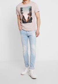 T-shirt rosa chiaro a maniche corte con grafica di una palma, abbinato a jeans chiari strappati e sneakers bianche.