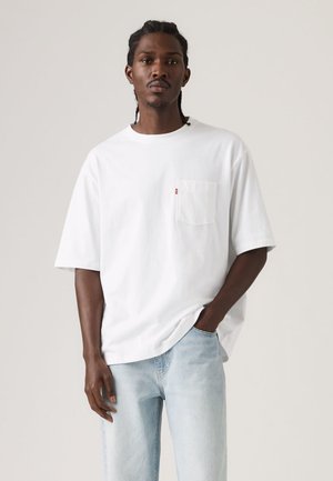 Fehér, rövid ujjú pamut póló, mellkasi zsebbel és egy kis piros logócímkével. Világoskék farmernadrággal párosítva.