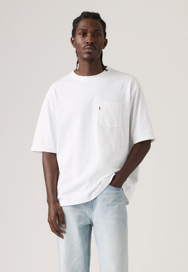 Λευκό κοντομάνικο βαμβακερό μπλουζάκι με τσέπη στο  στήθος και μια μικρή κόκκινη ετικέτα με λογότυπο. Συνδυάζεται με ανοιχτόχρωμα τζιν παντελόνια.