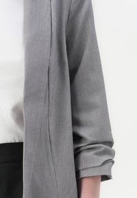 Cardigan gris à manches longues avec poignets retroussés, fabriqué en tissu texturé. Porté par-dessus un haut blanc, avec une ceinture noire visible.