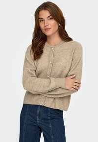 Beżowy kardigan z prążkowanej dzianiny, o okrągłym dekolcie i zapięciu na perłowe guziki. Długie rękawy, luźny krój i miękka tekstura. Noszony na niebieskie dżinsy.
