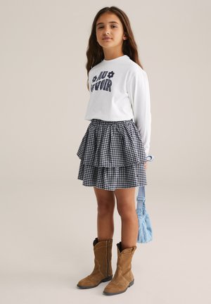 Chemise blanche à manches longues avec le texte "AU REVOIR", jupe à carreaux noir et blanc superposée, bottes marron et sac bleu clair.