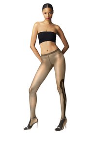 Donna in collant trasparenti beige con un motivo astratto nero sulla gamba destra, top bandeau nero e sandali con tacco alto argentati, in piedi con sicurezza.
