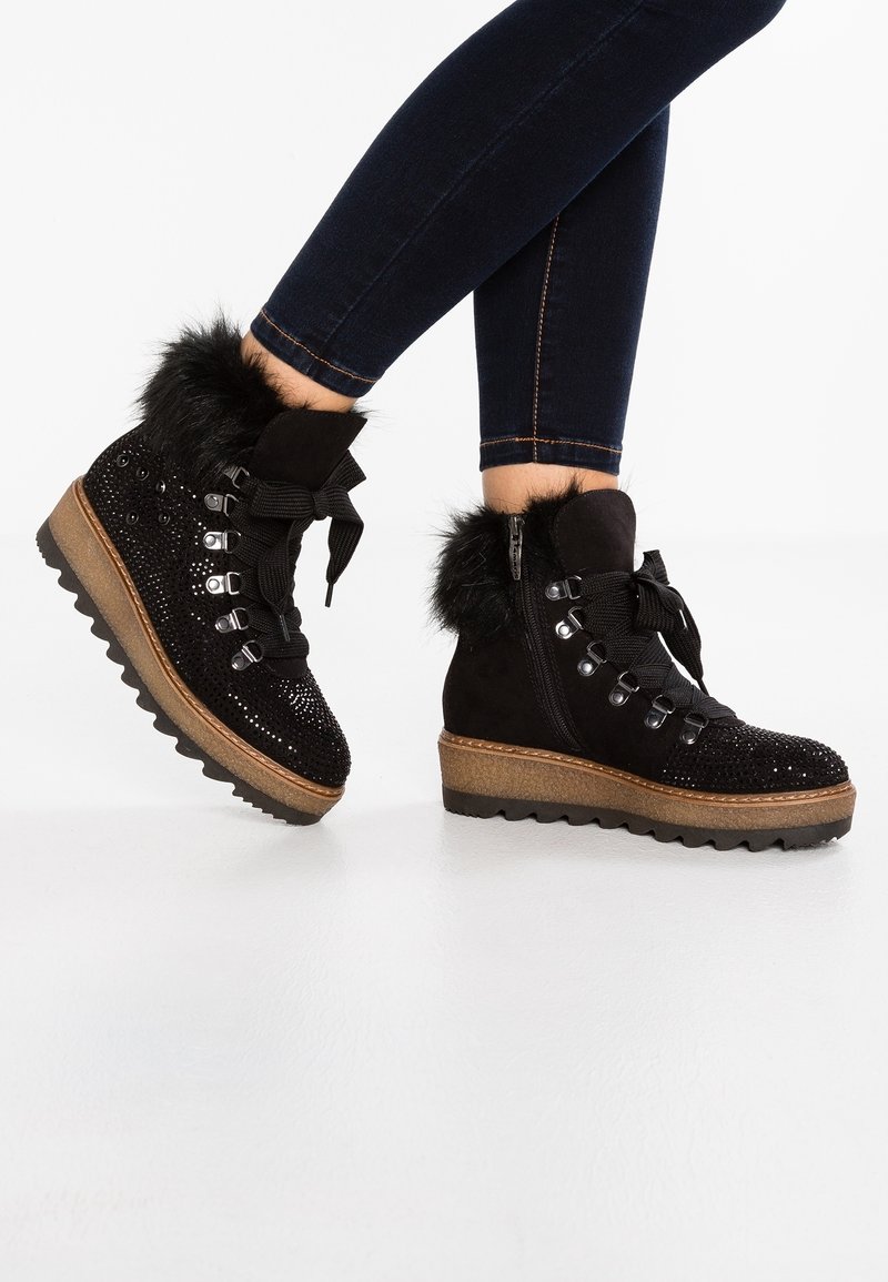 Bottes cheville noires bordées de fourrure avec des sequins, semelles épaisses marron, lacets noirs, portées avec un jean skinny foncé sur fond blanc.