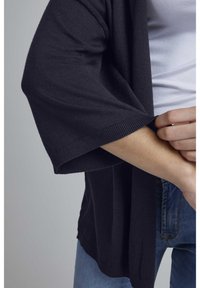 Svart stickad cardigan med vida ärmar, ribbade fållar och mjuk textur, i kombination med ljusa denimjeans. Nära bild av armen och plaggdetaljen.