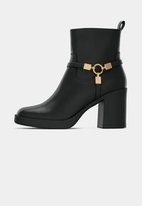 Crna faux kožna čizma do gležnja s kvadratnom blok petom, koja ima zlatnu opremu s remenom i kružnim detaljem.