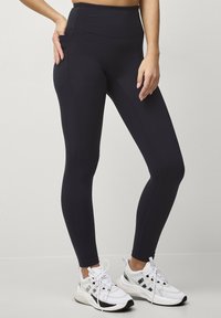 Højtaljede sorte leggings lavet af strækbart stof, med sidelommer, flade sømme og en tætsiddende pasform. Sammen med hvide kondisko.