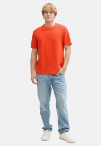 Rød t-shirt med korte ærmer i et tekstureret stof, parret med lyseblå denimjeans og hvide sneakers, stående foran en ensfarvet baggrund.