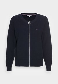 Námorní svetr pletený z vlny s zipovým zapínáním, širokými rukávy a žebrovanými manžetami. Obsahuje kruhový táhlo zipu a malé logo na hrudi.