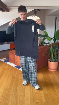 Mladenič drži novo črno majico z dolgimi rokavi z etiketami v dnevni sobi s lesenim tlakom in oranžno kavčem.