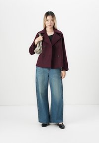 Ungdommelig kvinne iført en mørk lilla dobbelspent frakk, bredbeinede blå jeans, svarte leiligheter, og bærer en veske med slangeskinnmønster over skulderen.