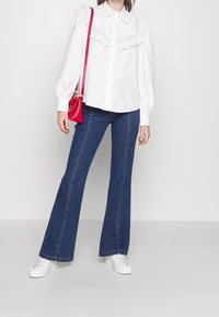 Camicia bianca con dettagli in pizzo sul colletto, abbinata a jeans svasati blu scuri e sneakers bianche. È visibile una borsa a tracolla rossa.