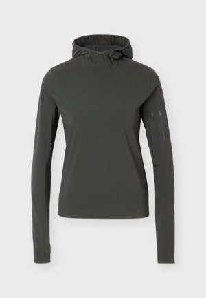 Sweat à capuche slim foncé avec trous pour les pouces, associé à des leggings taupe avec fentes aux chevilles, porté par une personne debout en baskets blanches.