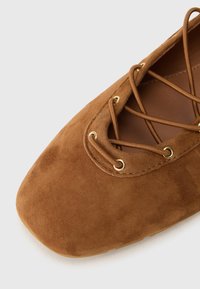Scarpa piatta in suede marrone con punta quadrata e lacci incrociati infilati attraverso occhielli dorati.