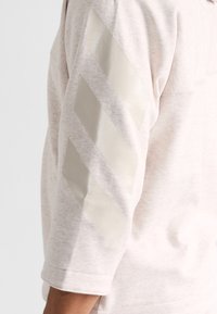 Sudadera beige claro con una textura suave. El codo presenta un patrón de chevrón gris claro y brillante, y las mangas son de longitud 3/4.