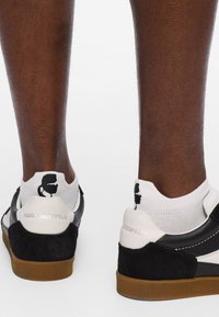 Zapatillas en blanco y negro con materiales de ante y cuero, que presentan una suela de goma contrastante y un logotipo distintivo en el talón y en los calcetines.