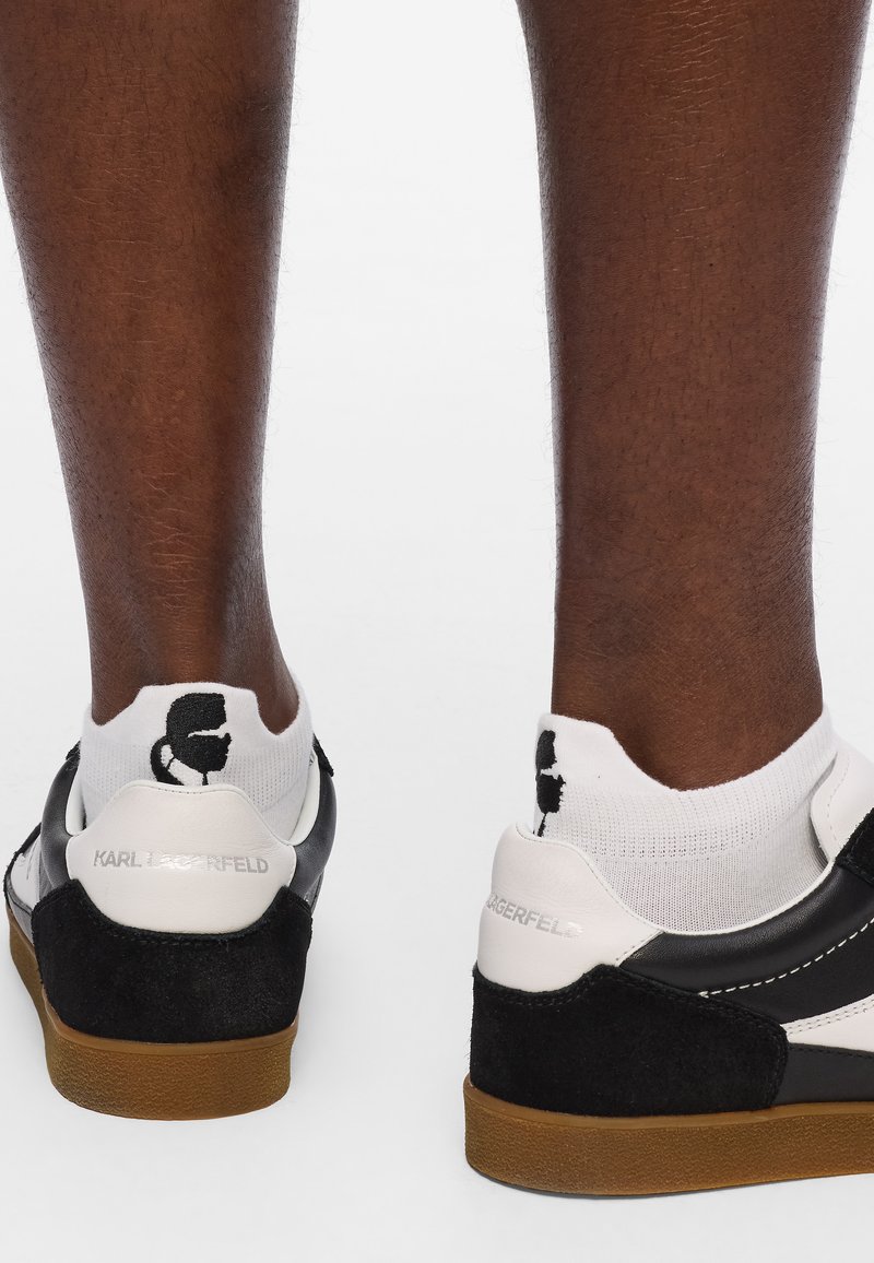 Zapatillas en blanco y negro con materiales de ante y cuero, que presentan una suela de goma contrastante y un logotipo distintivo en el talón y en los calcetines.