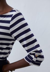 Marinblå och vitrandig långärmad topp med båtringning, utrustad med uppvikta ärmar och knappdetaljer nära manschetterna.