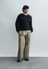 Pull en maille noire avec des motifs texturés, associé à un pantalon beige à jambes larges. La tenue est complétée par des baskets noires avec des bandes blanches.