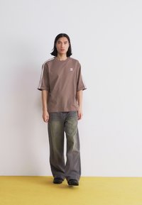 Ruda per didelį t-shirt'ą su baltomis šoninėmis juostomis ir logotipu, derinama su plačiais, išblukusiais žaliais džinsais ir juodais batais. Paprasta fono.