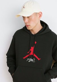 Hanorac negru cu un logo roșu Jordan și textul "Flight"; purtat sub o șapcă crem. Materialul este moale; designul este casual și sporty.
