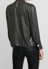 Blusa nera glitterata con maniche lunghe e apertura a chiave sulla schiena. Presenta bottoni ai polsini e una vestibilità ampia, realizzata in un tessuto lucido e texture.