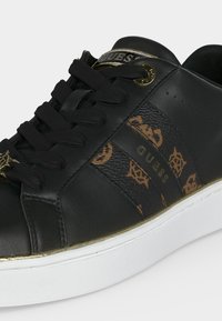 Baskets en cuir noir avec lacets noirs, panneau latéral marron à motif avec inscription "GUESS", accents dorés et semelle en caoutchouc blanche.
