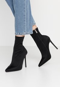 Stivaletti neri alla caviglia con una finitura satinata elegante, punta affusolata, alto tacco a stiletto e chiusura con zip laterale, abbinati a jeans cropped azzurri.