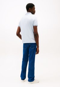 Világoskék galléros póló fekete-fehér szegéllyel, kék farmernadrággal és fehér tornacipővel, hátulnézetből.