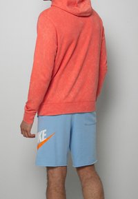 Felpa corallo con finitura testurizzata, abbinata a pantaloni corti azzurri chiari con un audace logo Nike bianco e dettaglio dello swoosh arancione.