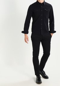 Černá džínová košile s dlouhým rukávem a dvěma náprsními kapsami, spárovaná s úzkými černými kalhotami a černými šněrovacími botami.