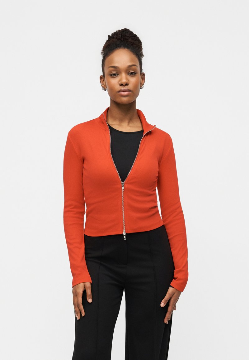 Κόκκινο μπουφάν με φερμουάρ, μακριά μανίκια, ριμπ ύφασμα και κοντή εφαρμογή. Συνδυασμένο με μαύρο τοπ και μαύρο παντελόνι.