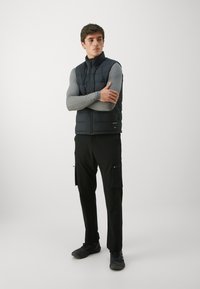 Crna pufasta prsluk s visokim ovratnikom, nošen preko svijetlosive košulje dugih rukava. Crne cargo hlače s bočnim džepovima i tamnim cipelama.