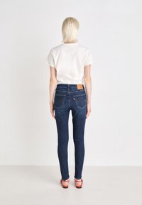 Schlanke blaue Denim-Jeans mit mittelhohem Bund, Gesäßtaschen, gelben Nähten und einem Leder-Logo-Patch. Begleitet von einem schlichten weißen T-Shirt.