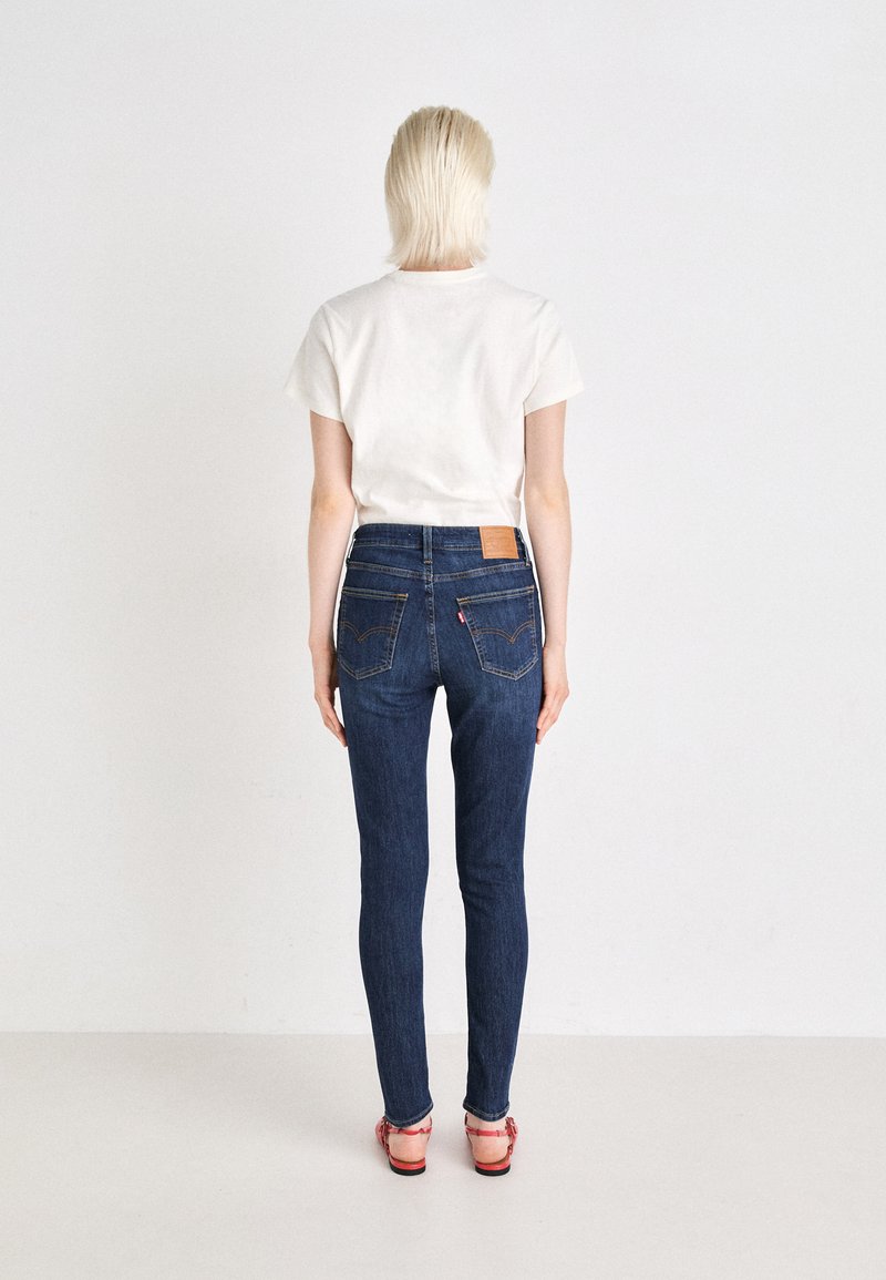 Schlanke blaue Denim-Jeans mit mittelhohem Bund, Gesäßtaschen, gelben Nähten und einem Leder-Logo-Patch. Begleitet von einem schlichten weißen T-Shirt.
