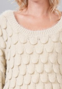 Cream-colored sweater with a textured design featuring overlapping scalloped patterns. Knit material with a wide neckline and soft finish.