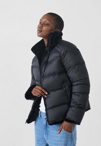 Giubbotto imbottito nero con colletto alto, chiusura con zip e dettagli in pelliccia sintetica. Design trapuntato realizzato in materiale leggermente lucido, abbinato a jeans azzurri chiari.