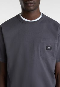 Camiseta gris de manga corta hecha de algodón, con cuello redondo, que presenta un bolsillo en el pecho y una etiqueta de marca rectangular negra.