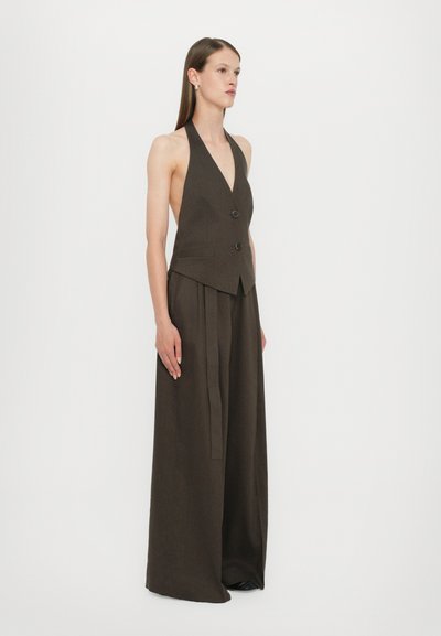 Halterneck-Weste aus dunkelbraunem Stoff mit zwei vorderen Knöpfen, kombiniert mit weiten Hosen, die ein seitliches Bindedetail und eine glatte Textur aufweisen.