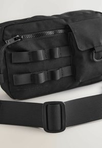 Black fabric bag with front zip pocket, vertical webbing, and loop accents. Includes a matching adjustable strap with a plastic buckle.