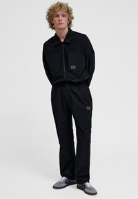 Zwart tweedelig outfit van gladde stof, met een ritsjack met twee grote zakken en een broek met elastische tailleband. Inclusief grijze sneakers.