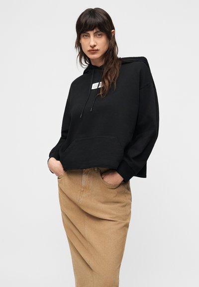 Άτομο με μακριά σκοτεινά μαλλιά, ντυμένο με μαύρη φούτερ μπλούζα με κουκούλα και καφέ φούστα, στέκεται με τα χέρια στις τσέπες μπροστά σε απλό φωτεινό φόντο.