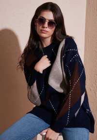 Navy blue knitted sweater with diamond patterns in brown and beige, featuring ribbed cuffs. Model wears round sunglasses and light blue jeans.