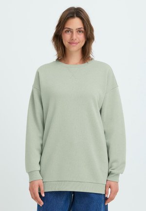 Jasnozielony, oversize'owy sweter z długimi rękawami, okrągłym dekoltem i ściągaczami na rękawach. Miękka tekstura, zestawiony z niebieskimi dżinsami.